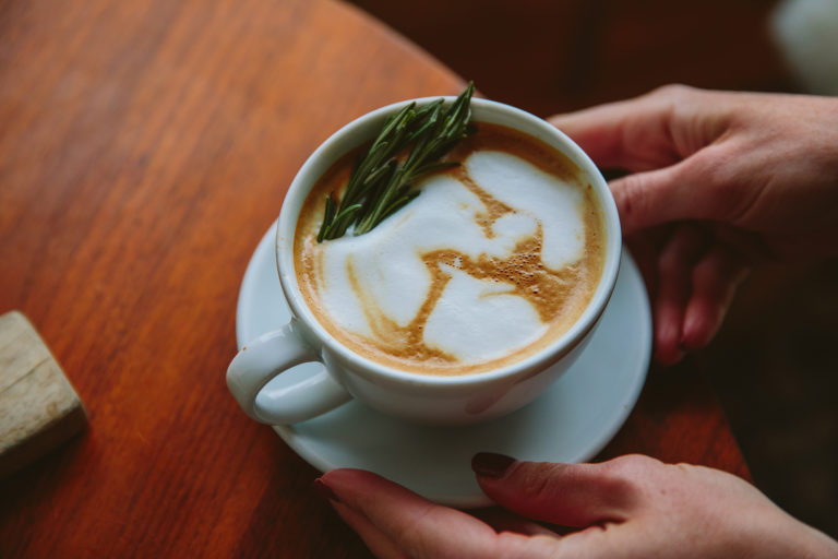 Hands holding a white cup and saucer with foamy coffee and a rosemary sprig on a wooden table.